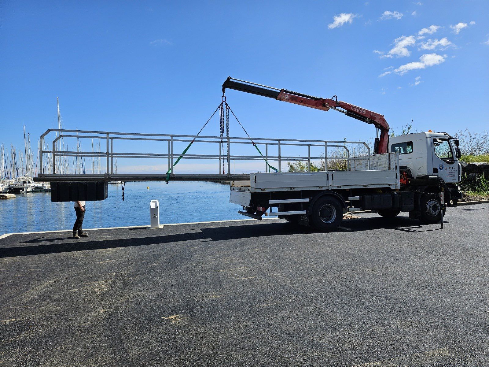 Une grue sur camion soulève une structure de garde-corps métallique près d'un plan d'eau
