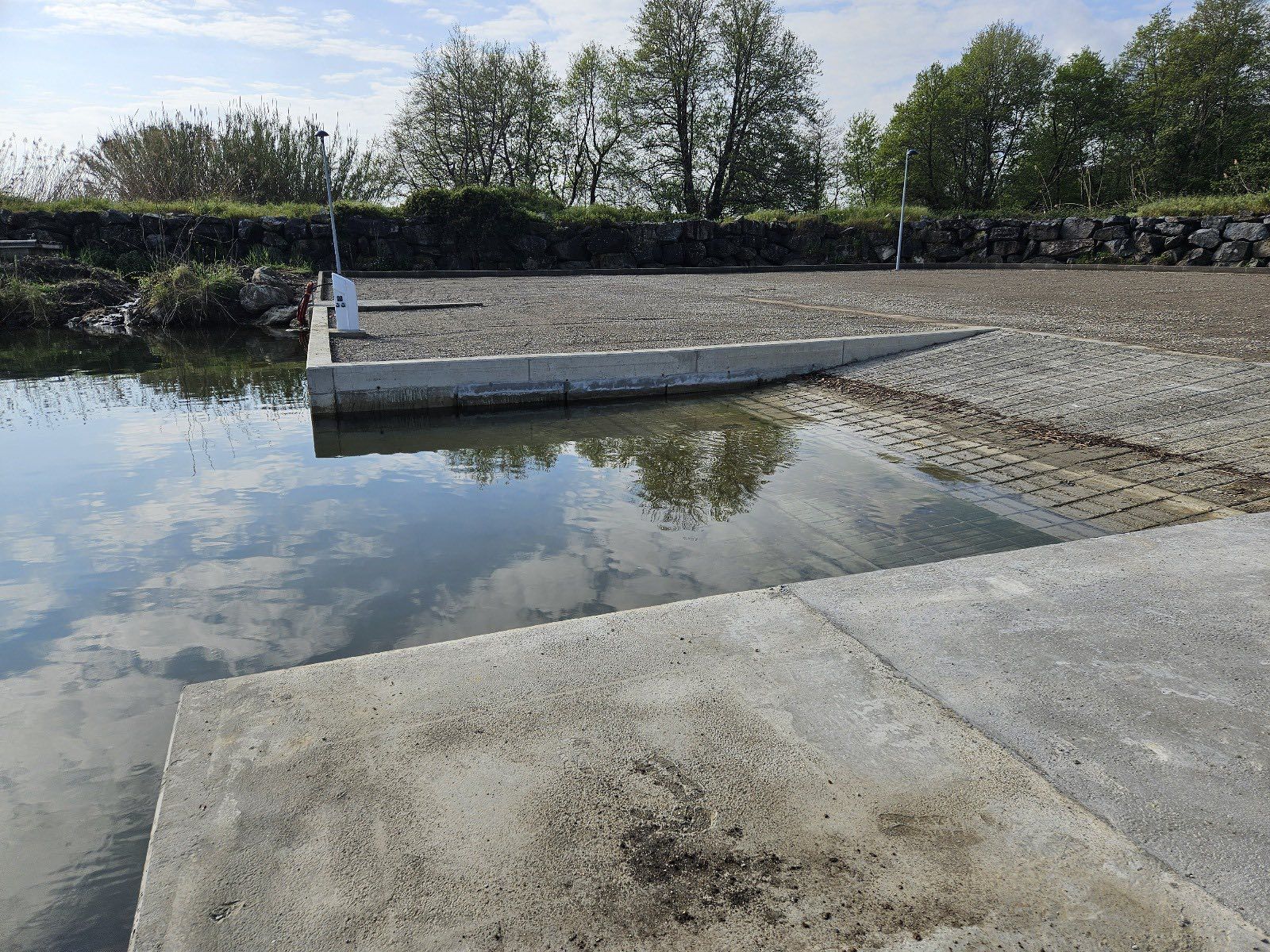 Rampe de mise à l'eau en béton, parking en gravier, arbres en arrière-plan, ciel nuageux