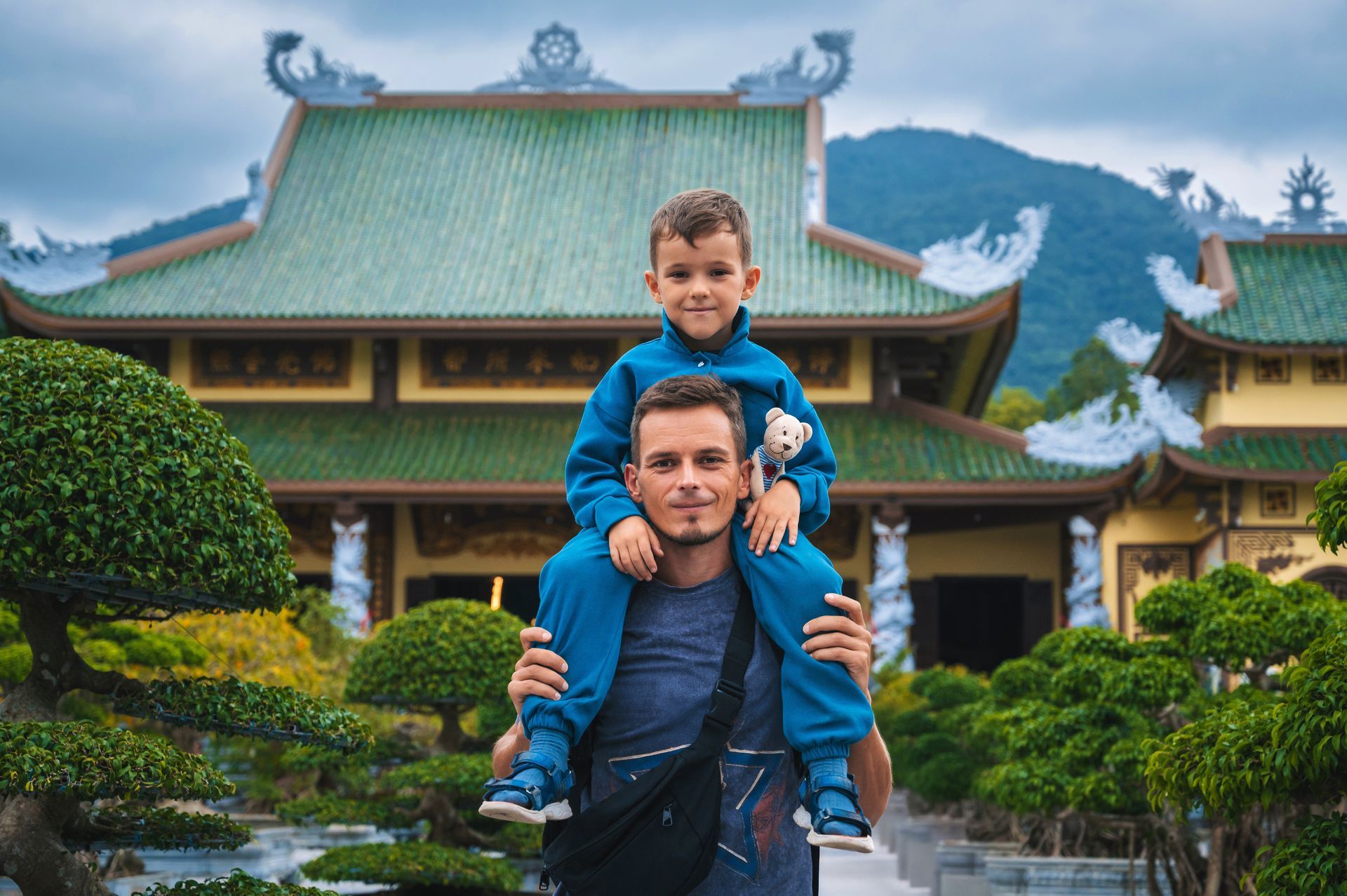 Un homme portant un enfant souriant sur ses épaules devant un temple japonais au toit vert.