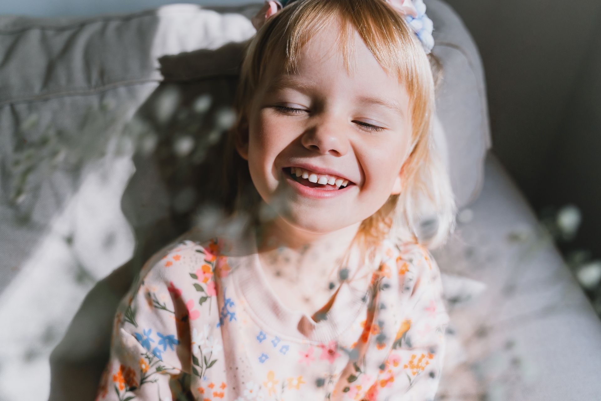 Enfant souriante est assise sur son lit, les yeux fermés. Le soleil éclaire son visage.