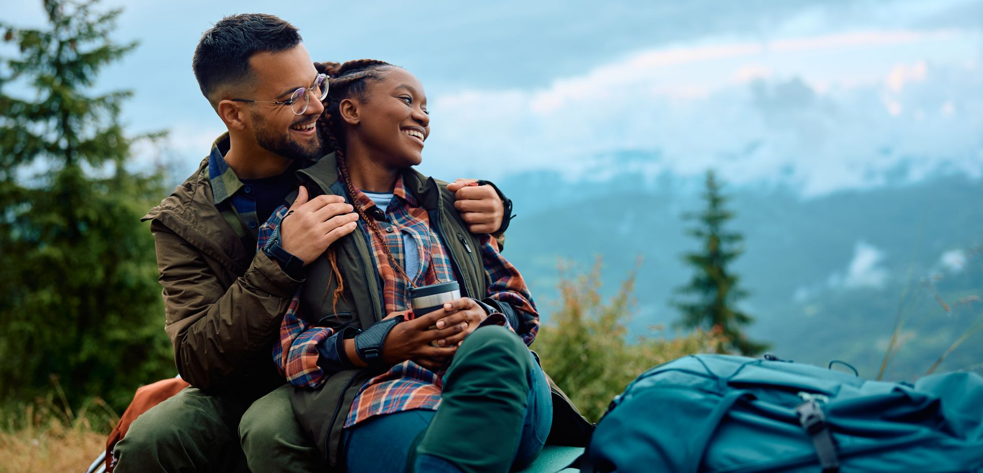 Un couple assis sur un promontoire montagneux, souriant et enlacé, avec leurs sacs à dos.