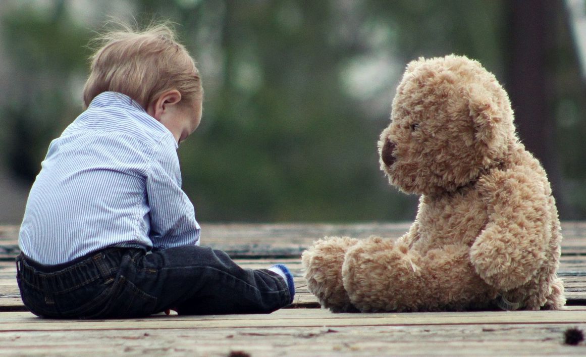 Enfant assis sur une terrasse en bois face à un ours en peluche.