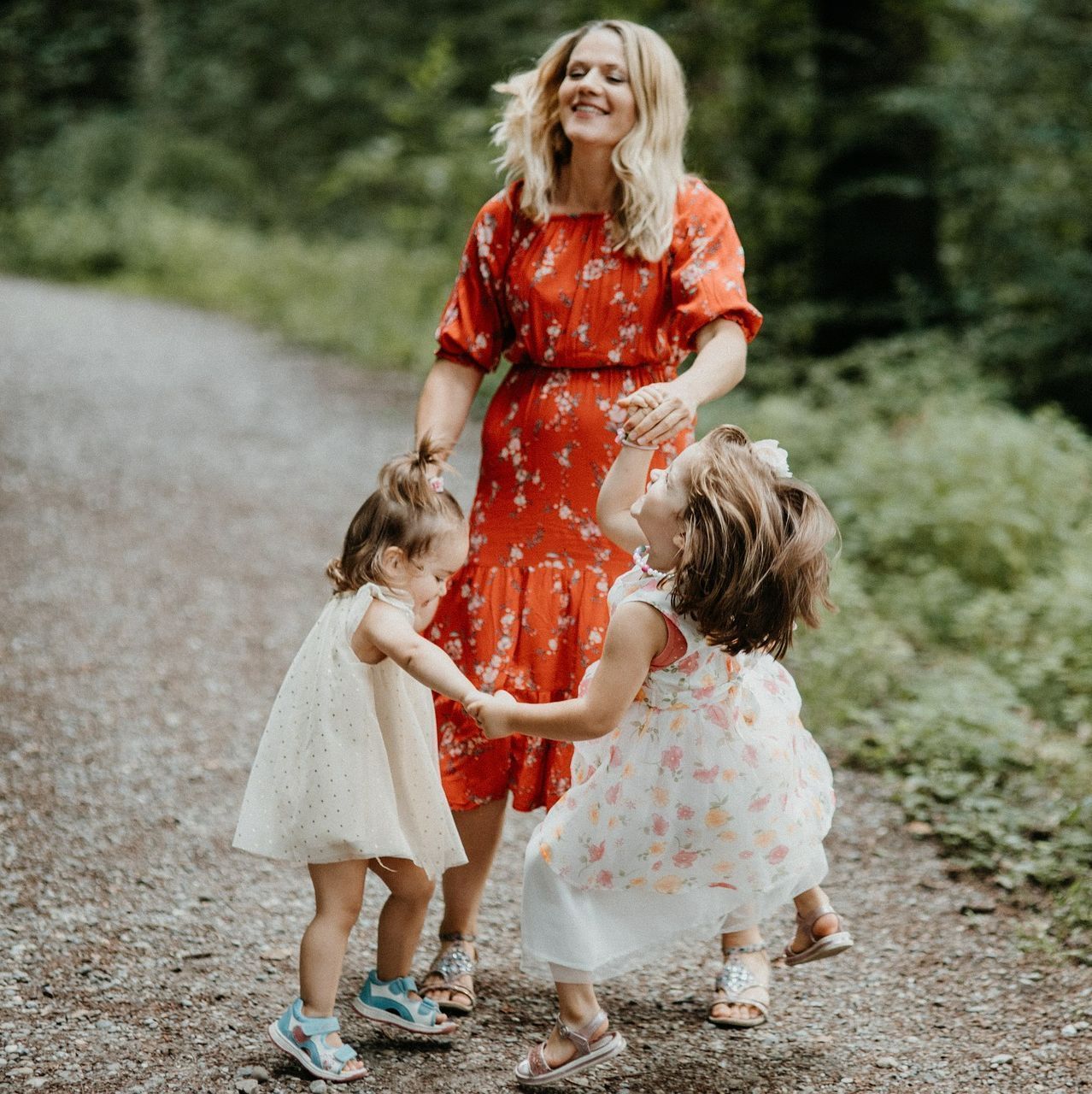 Une femme en robe rouge tenant la main de deux enfants sur un chemin de gravier dans un parc verdoyant.