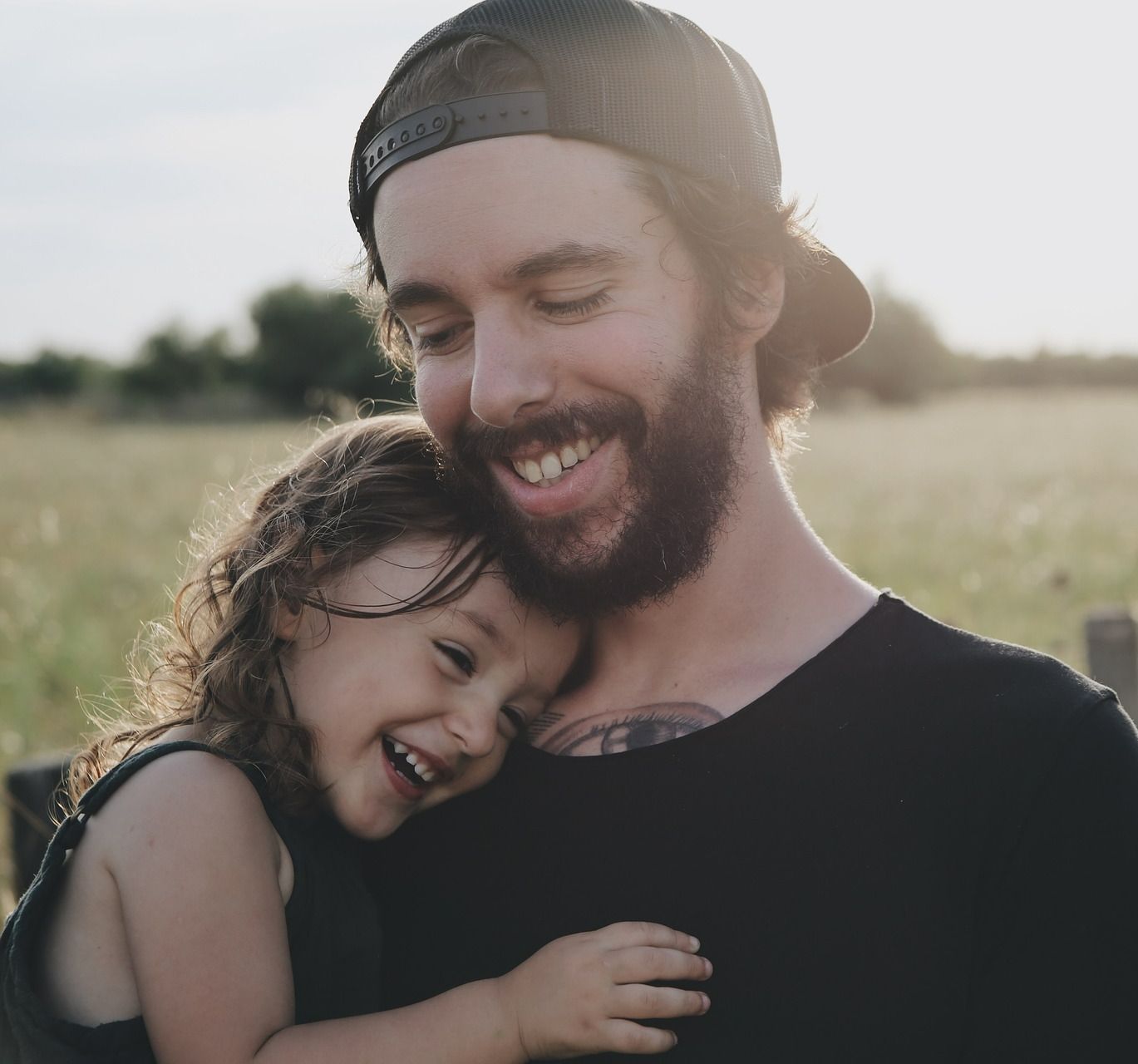 Un homme souriant, casquette à l'envers, enlace un enfant rieur dans un champ ensoleillé.