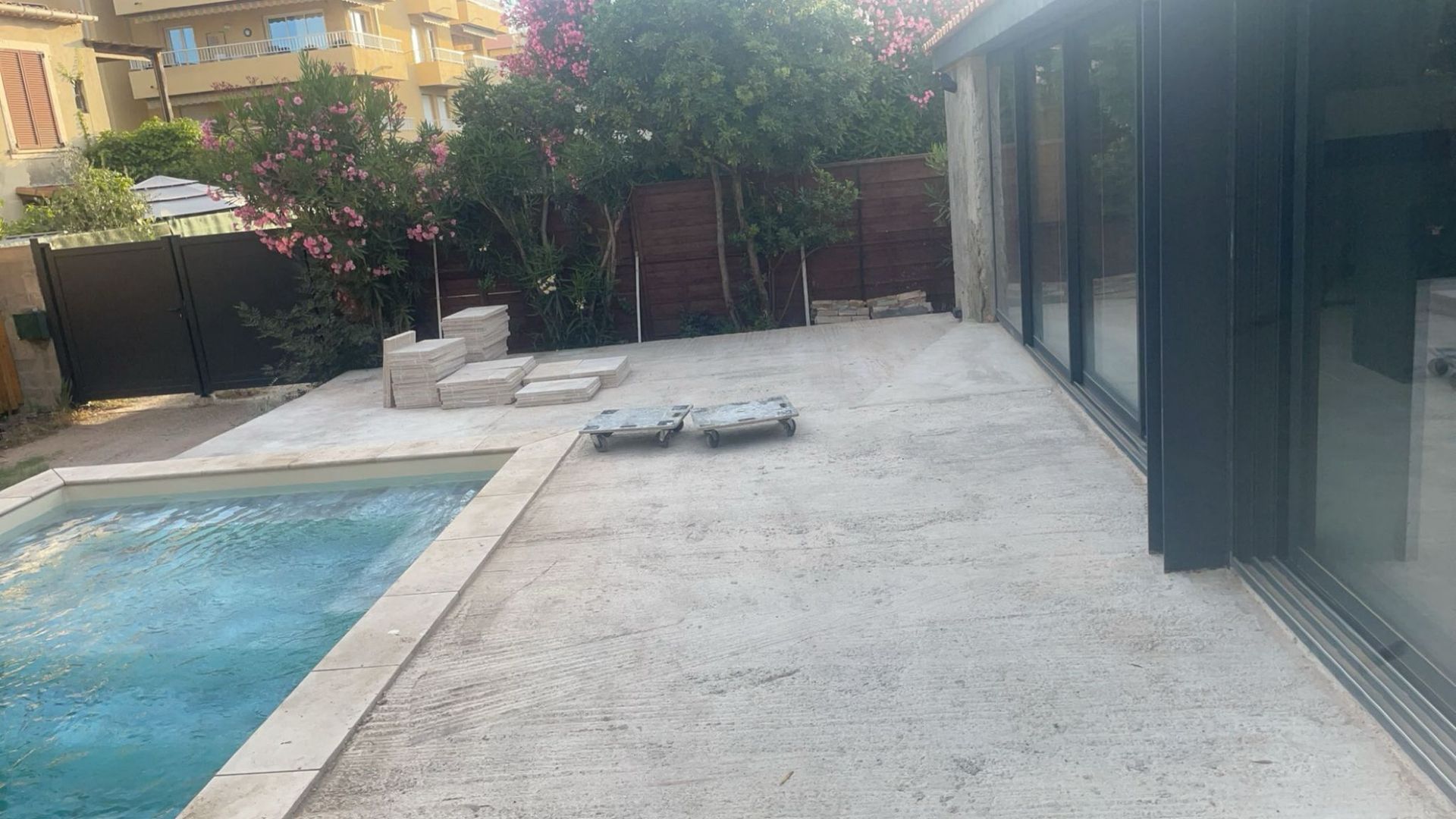 Un jardin avec piscine et terrasse. La terrasse est en béton et jouxte un bâtiment aux portes vitrées.