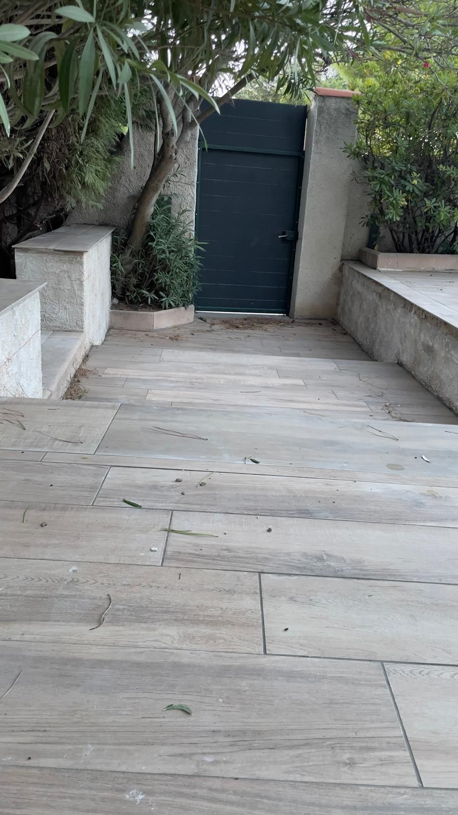 Un chemin de pierre mène à un portail en bois sombre, flanqué de murs en béton et de verdure.