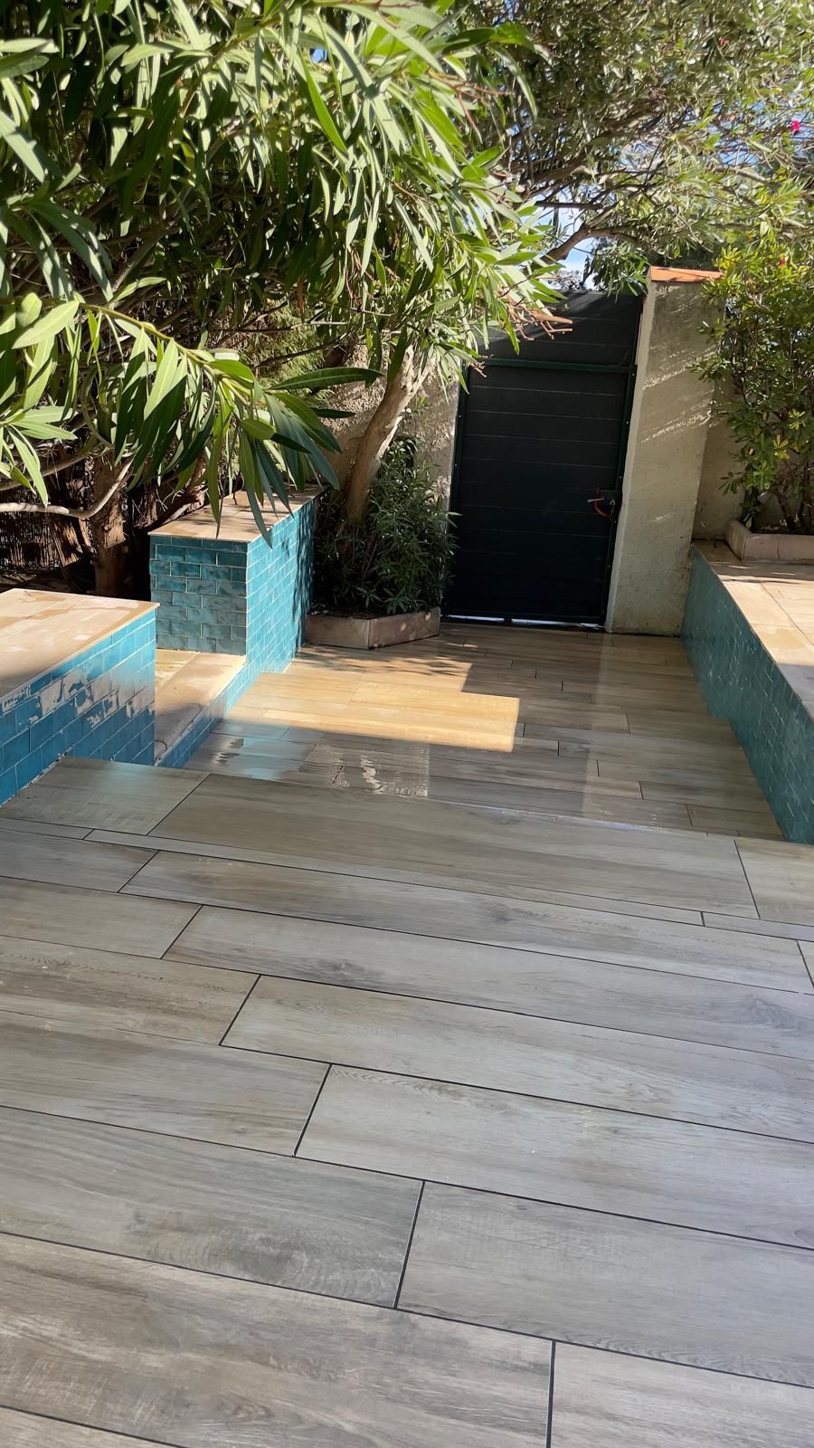 Une terrasse en pierre bordée de carreaux bleus mène à un portail noir, avec une végétation luxuriante en arrière-plan.