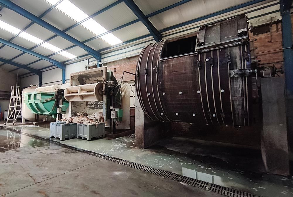 Gran máquina industrial dentro de un almacén, con un tambor cilíndrico y tuberías bajo un techo metálico.