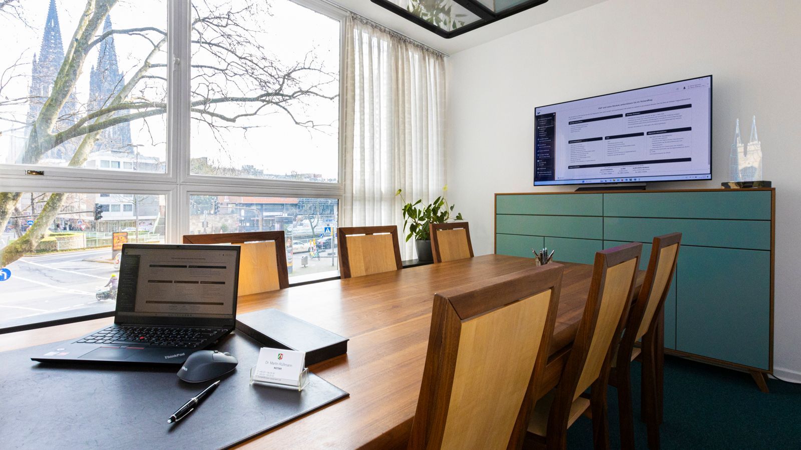 Modernes Heimbüro mit Holztisch, Laptop, Tablet, Stühlen und einem türkisfarbenen Schrank an einem hellen Fenster
