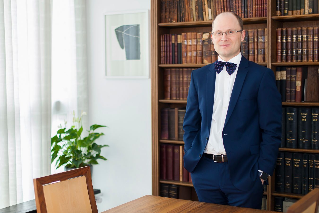 Mann im blauen Anzug steht an einem Holzschreibtisch in einem mit Büchern gefüllten Büro