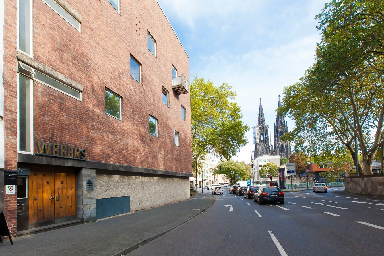 Straßenansicht mit Backsteingebäude, geparkten Autos, Bäumen und Kirchtürmen in der Ferne