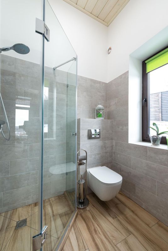 Salle de bains moderne avec douche vitrée, toilettes suspendues, carrelage gris et sol imitation bois près d’une fenêtre