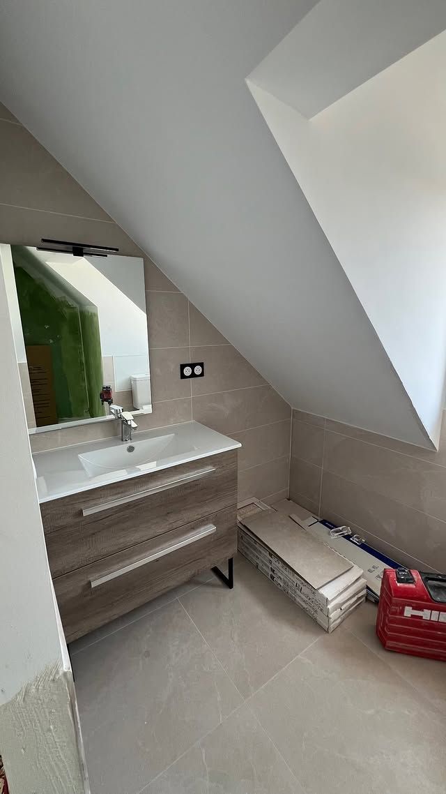 Salle de bains sous les combles avec plafond mansardé, miroir au-dessus du lavabo, meuble blanc et cartons au sol