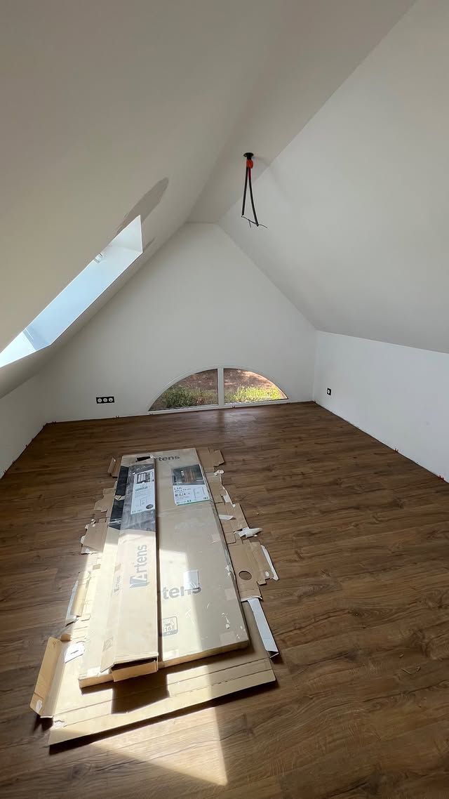 Pièce sous les combles inachevée, avec des murs blancs en pente, une fenêtre de toit, un parquet en bois et des planches empilées au centre.