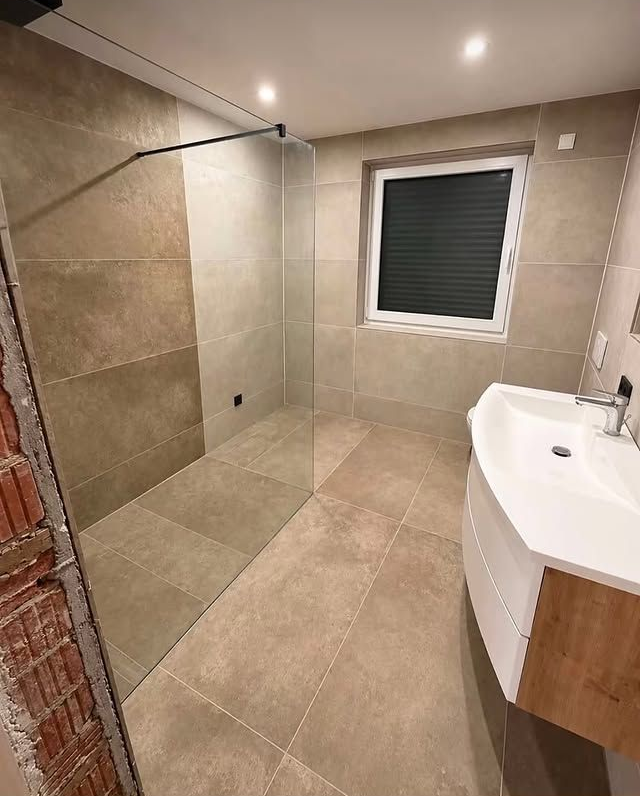 Salle de bains moderne carrelée avec douche à l’italienne, lavabo sur meuble et fenêtre avec store fermé.