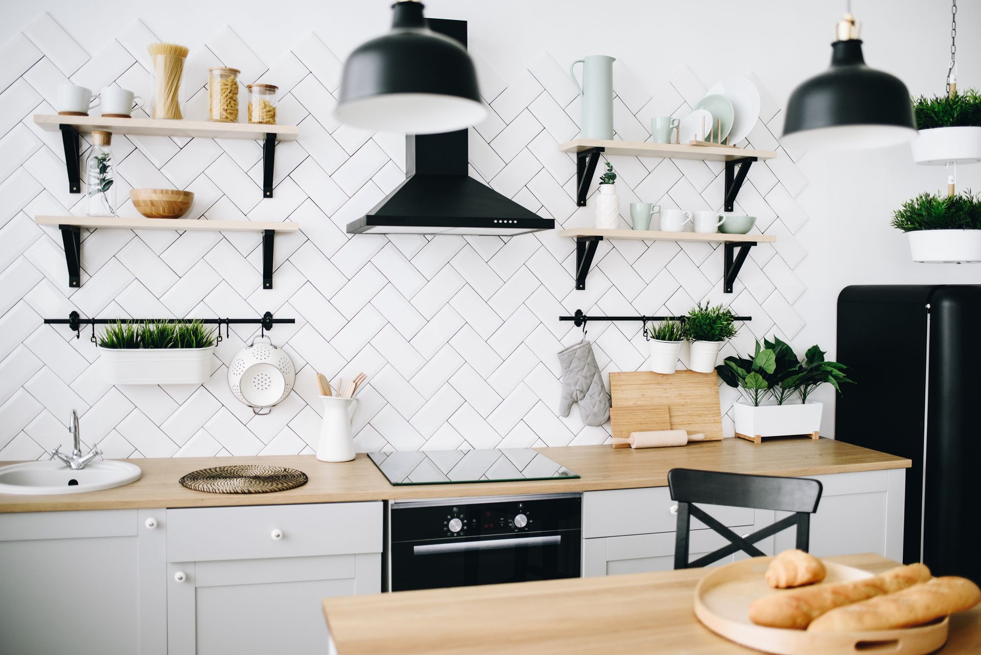 Cuisine moderne et lumineuse avec crédence en carreaux blancs, étagères ouvertes, suspensions noires et table en bois avec du pain