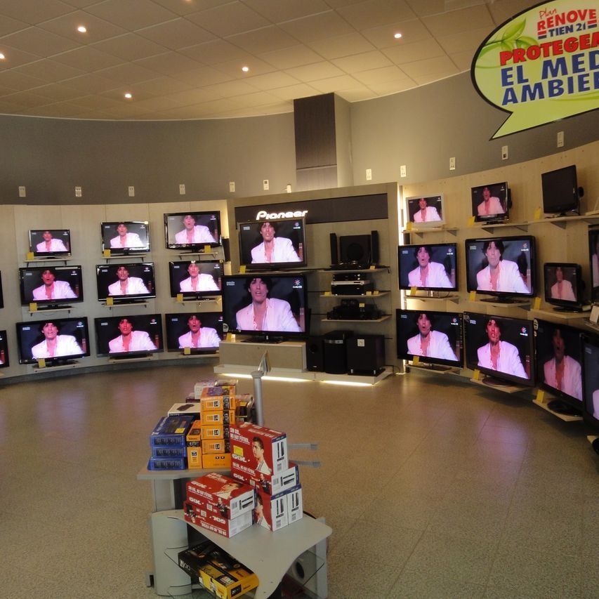 Dentro de una tienda, una exhibición de televisores con panel de la marca Pioneer, y estante de productos en primer plano.