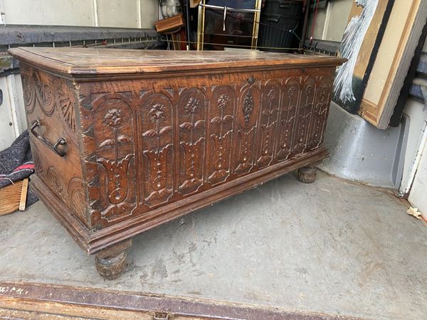 Un coffre ancien en bois orné de sculptures florales complexes sur le devant et reposant sur des pieds tournés sombres, posé sur le plancher d'un camion.