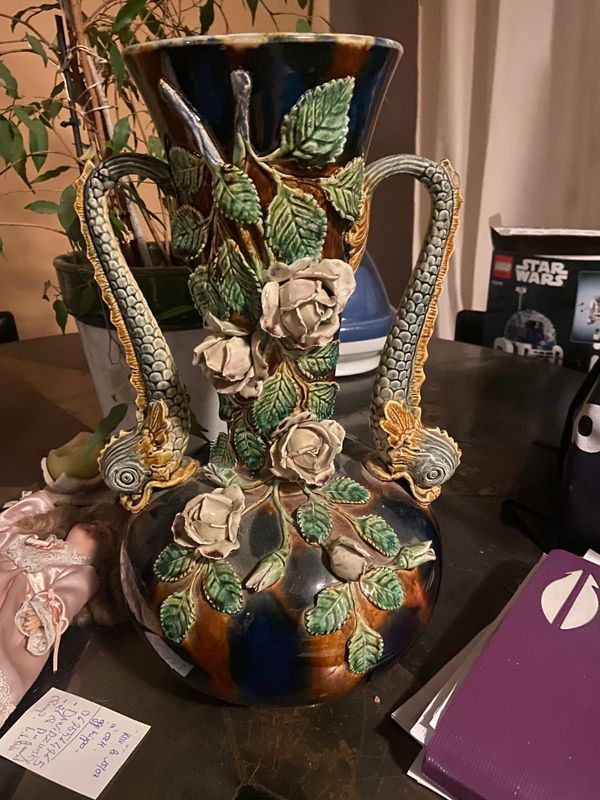 Vase en céramique orné de sculptures de roses couleur crème, de feuilles vertes et de poignées décoratives texturées, posé sur une table.