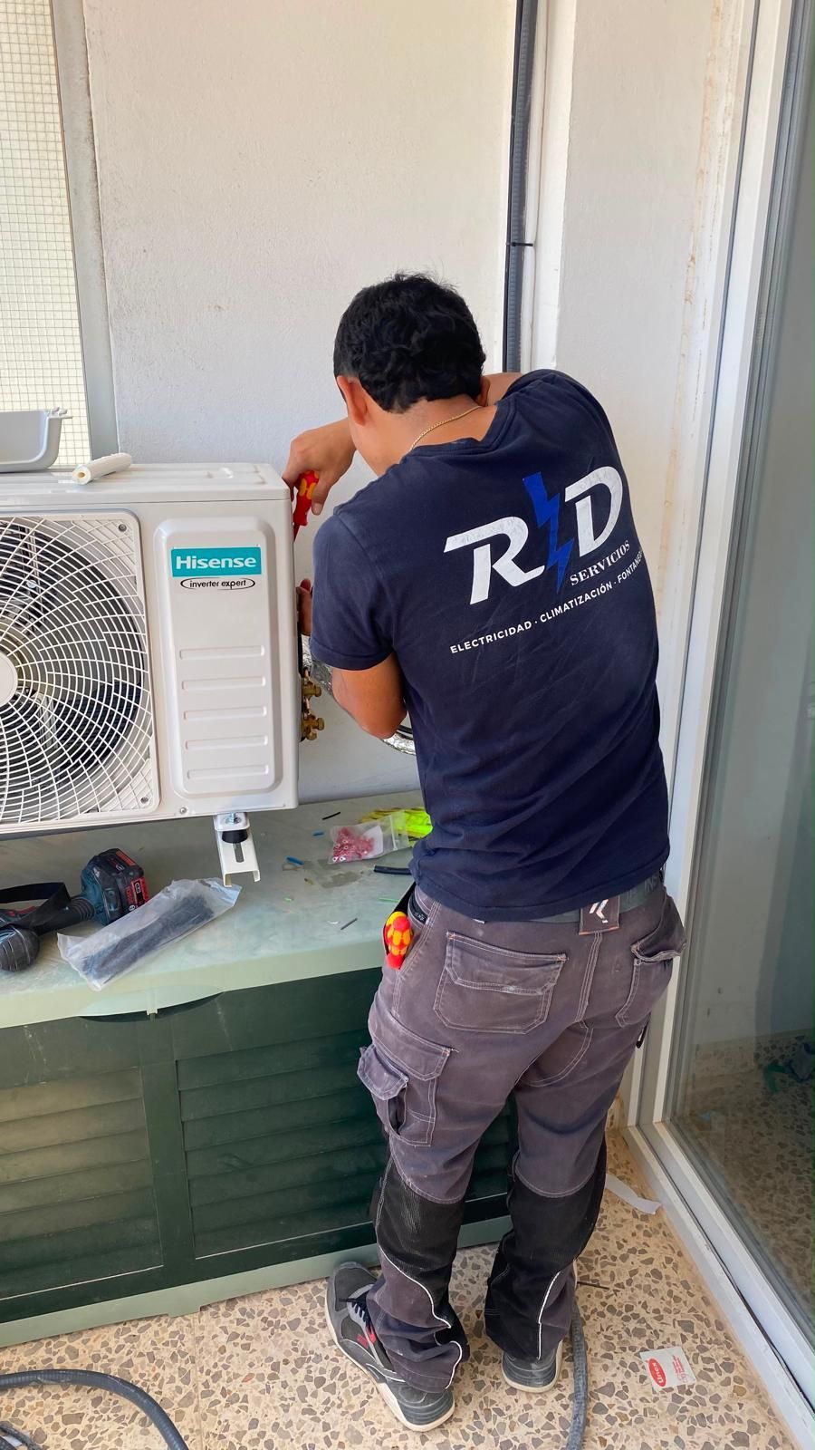 Técnico reparando una unidad de aire acondicionado exterior junto a una pared y una puerta abierta.