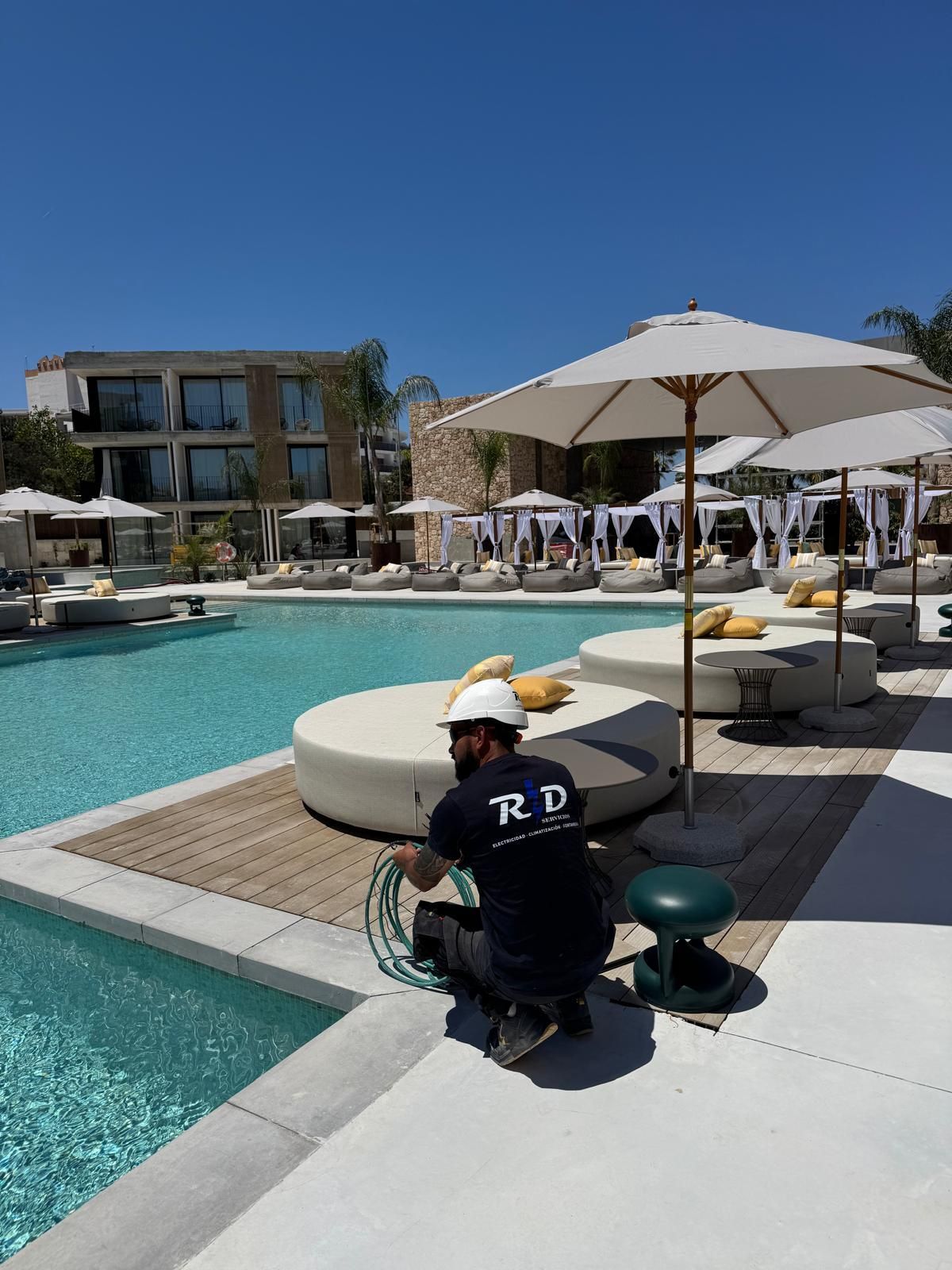 Un trabajador arrodillado junto a la piscina de un complejo turístico, ajustando mangueras cerca de sombrillas blancas