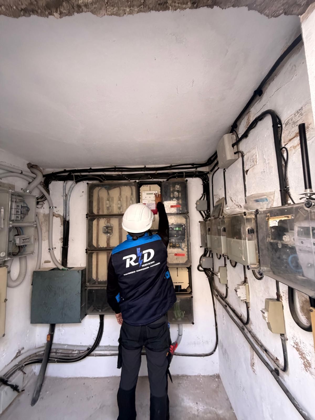 Técnico inspeccionando paneles eléctricos en un cuarto de servicio estrecho, con casco y chaqueta azul.
