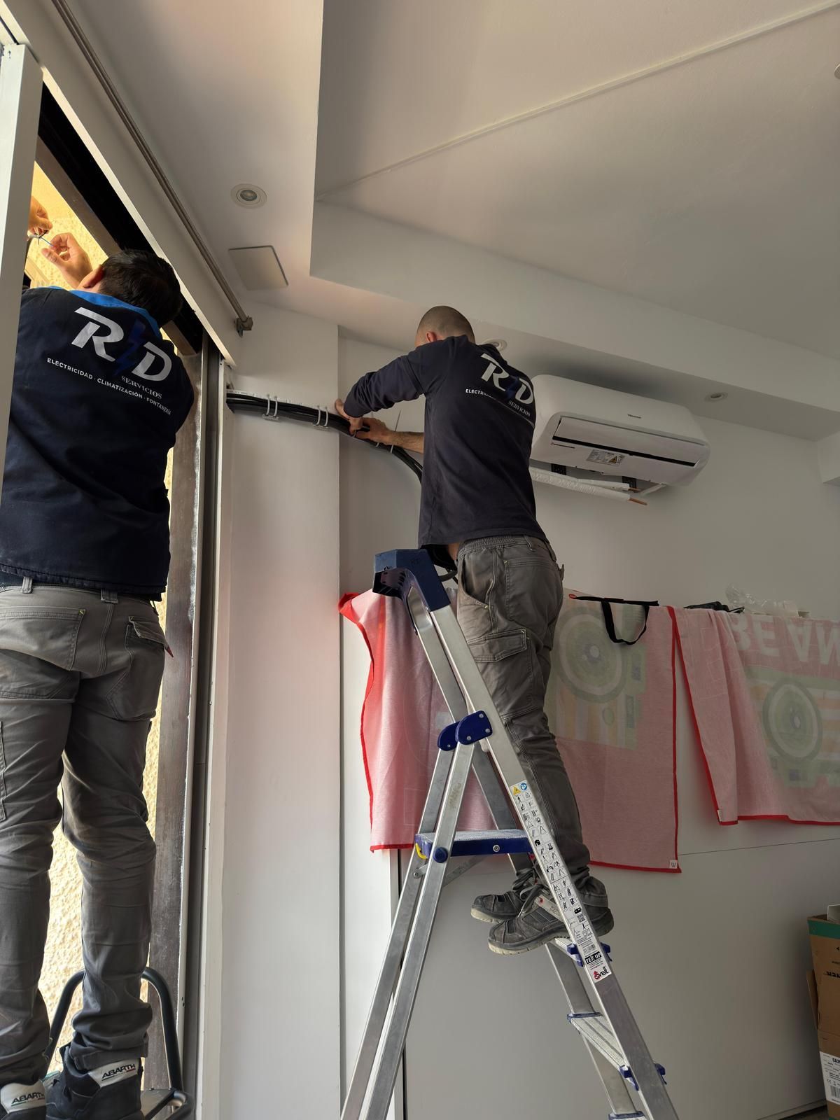 Dos trabajadores subidos a escaleras instalan un marco de ventana dentro de una habitación.