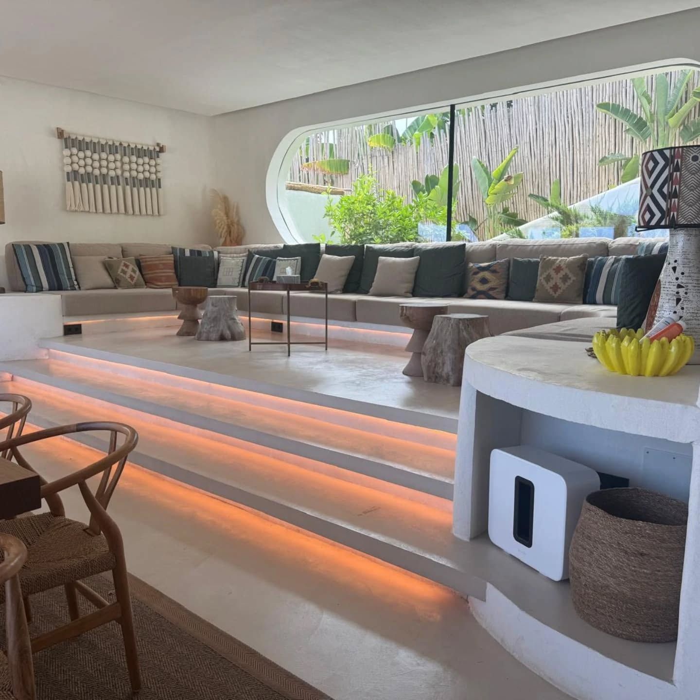 Salón moderno con sofá curvo, escalones iluminados, mesas bajas y grandes ventanales con vistas a la vegetación.