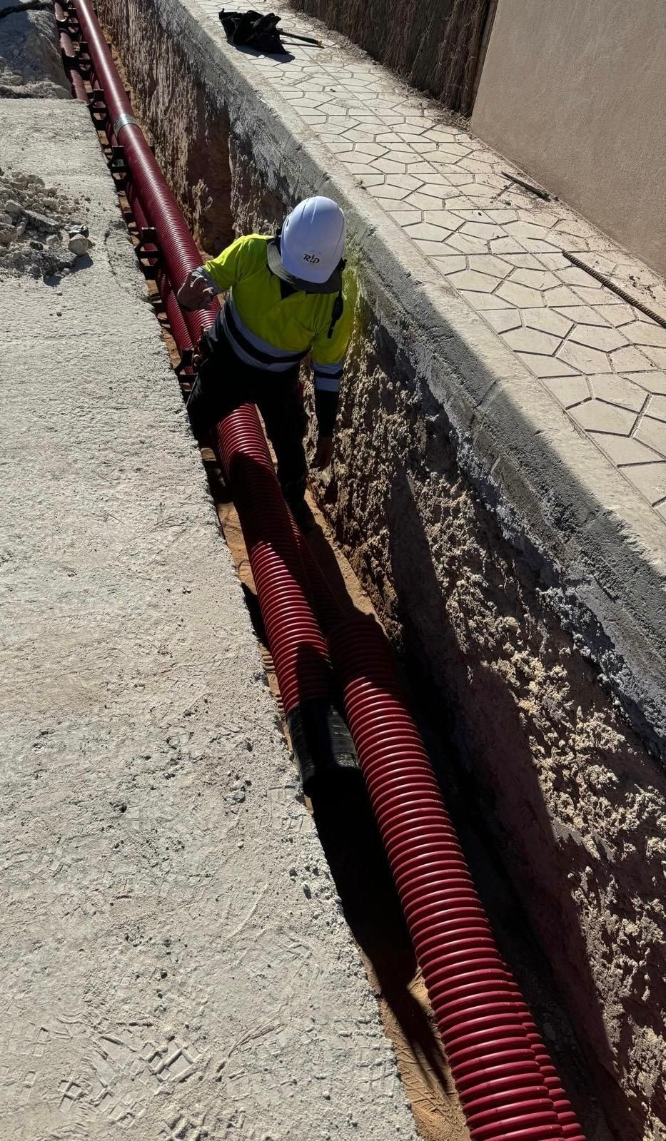 Obrero con casco junto a una zanja, manipulando conductos corrugados rojos en una zona de construcción.