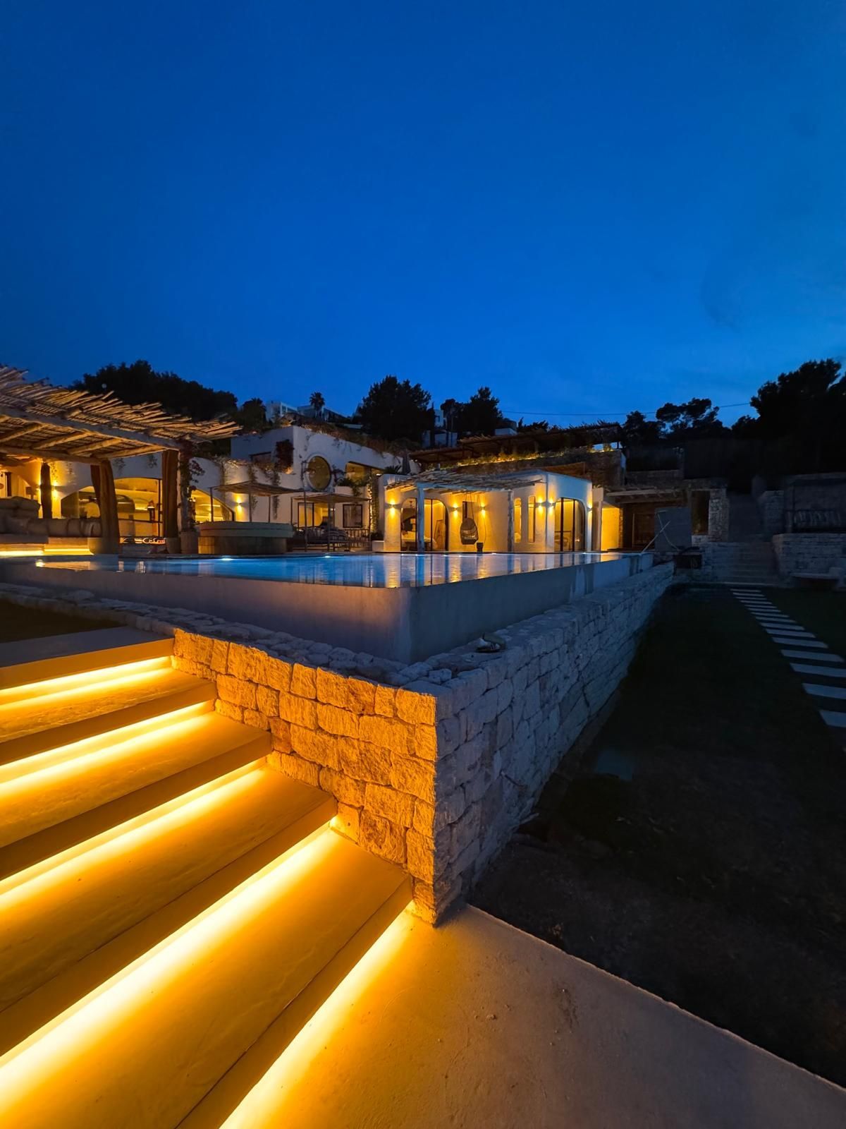 Patio de villa moderna al atardecer con escalones iluminados, terraza de piedra y cielo azul vespertino.