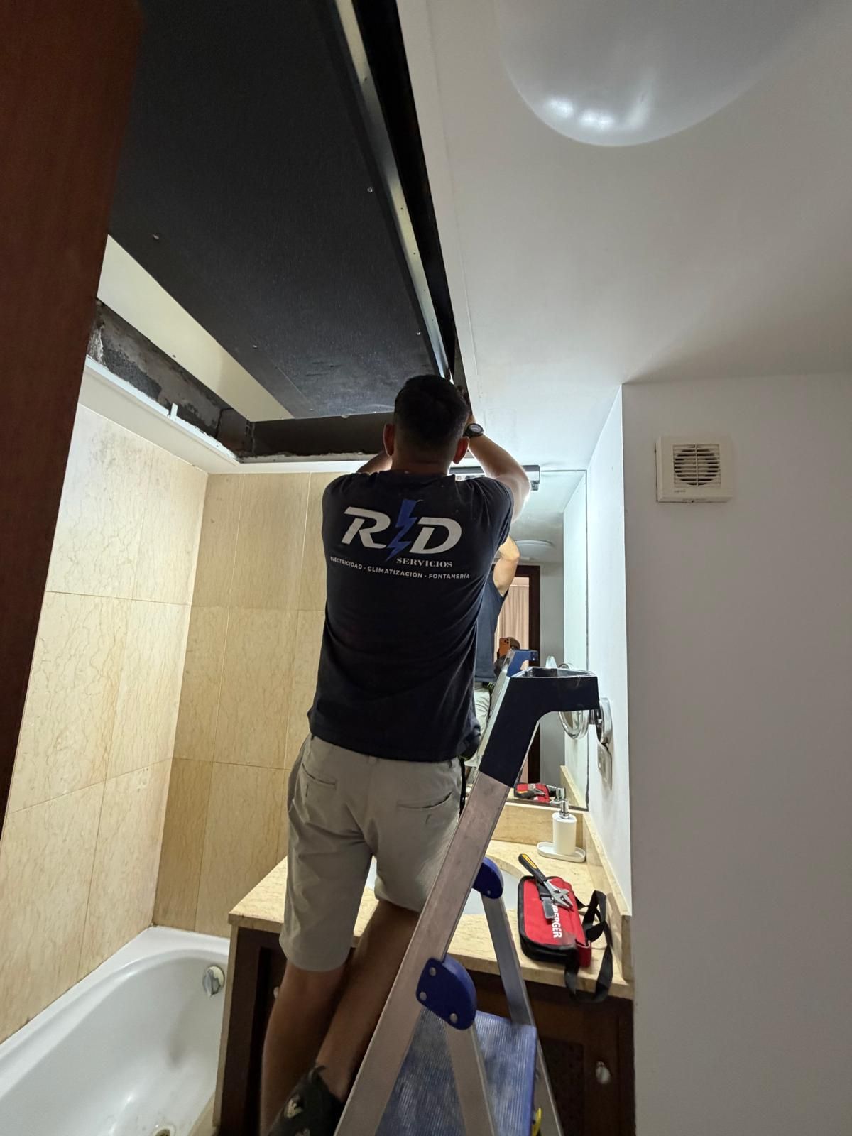 Persona de pie sobre una escalera que alcanza el techo en un pasillo estrecho, vestida con una camisa azul marino