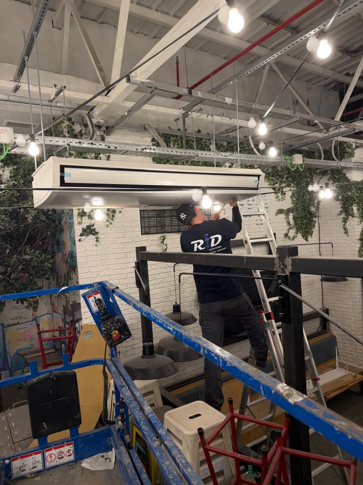 Zona de construcción interior con andamios, luces colgantes y trabajadores cerca de un muro de piedra y plantas.