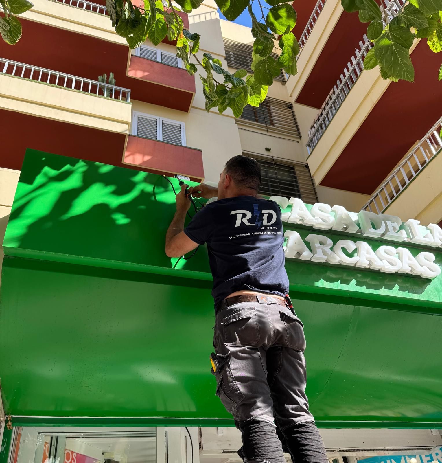 Una persona sube a una escalera y ajusta un letrero verde de una tienda debajo de balcones y hojas colgantes.