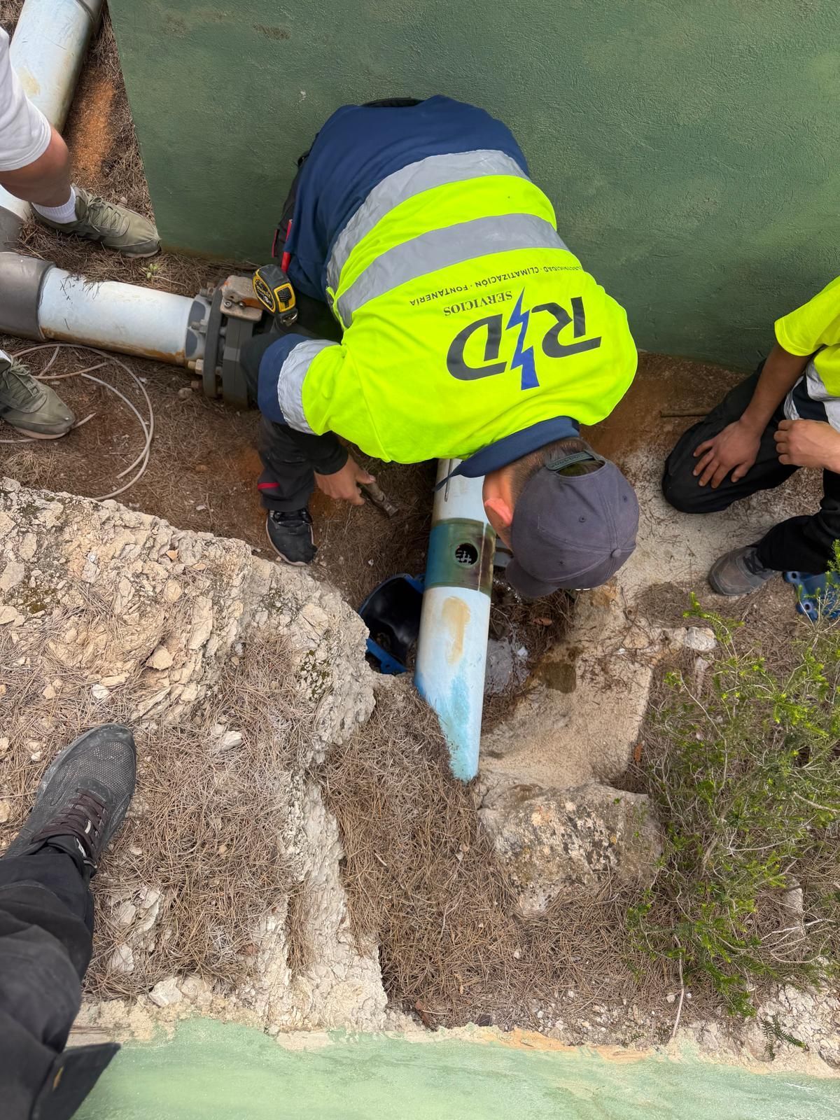 Un trabajador con chaleco reflectante repara una tubería al borde de una zanja fangosa.