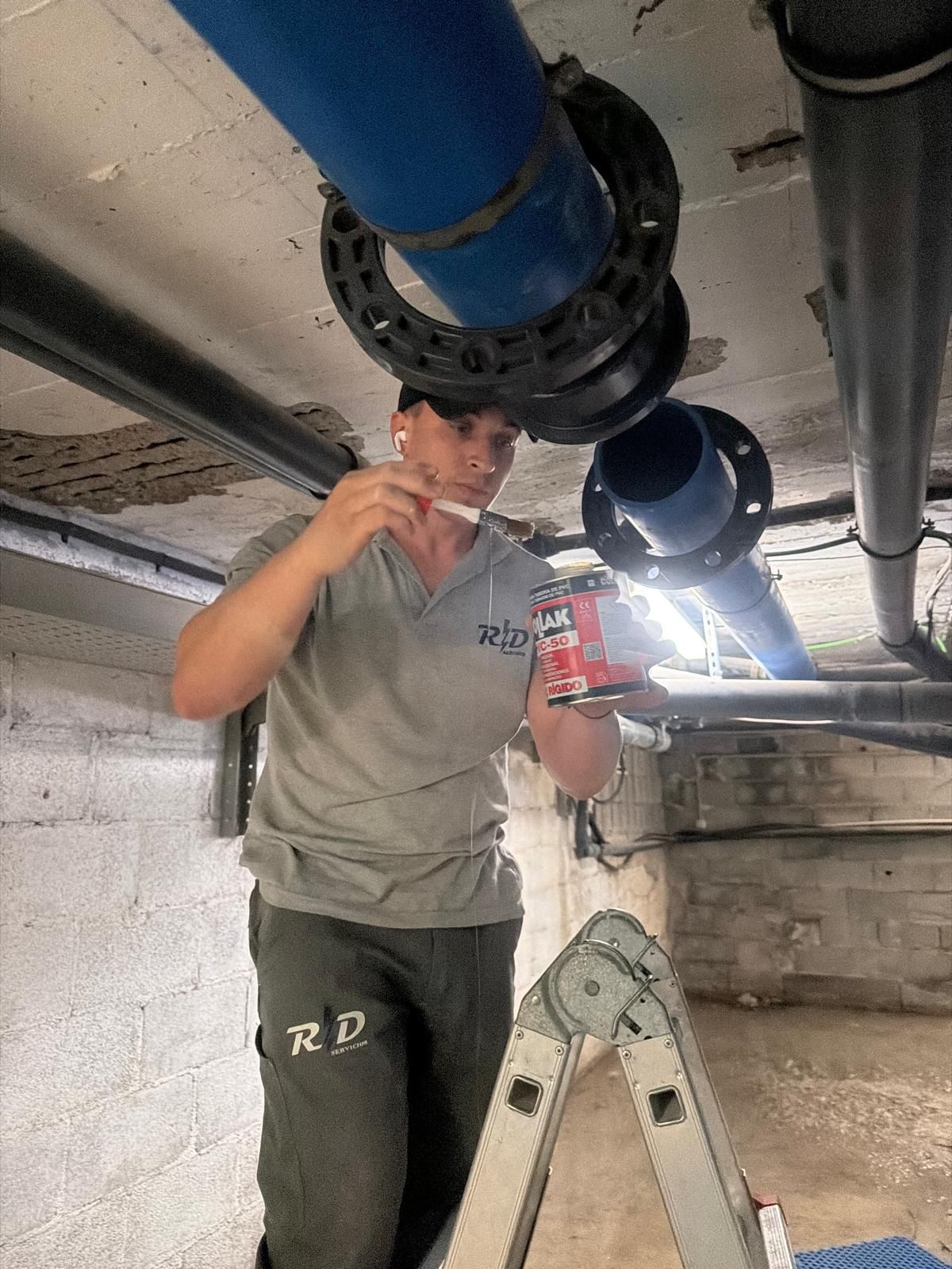 Un trabajador, subido a una escalera, inspecciona las tuberías azules y negras que cuelgan del techo