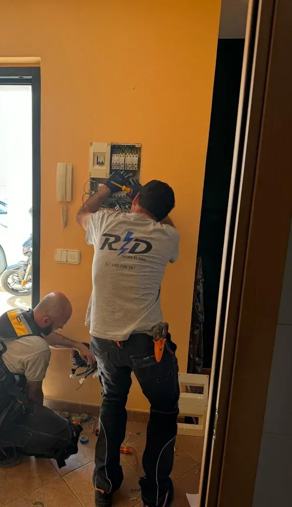 Dos trabajadores instalando una lámpara de pared en un pasillo con paredes naranjas y una puerta abierta.