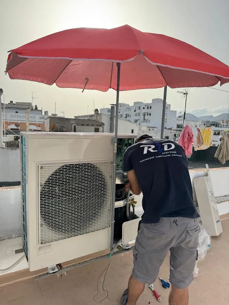 Persona trabajando en un aire acondicionado en la azotea bajo un paraguas rojo.