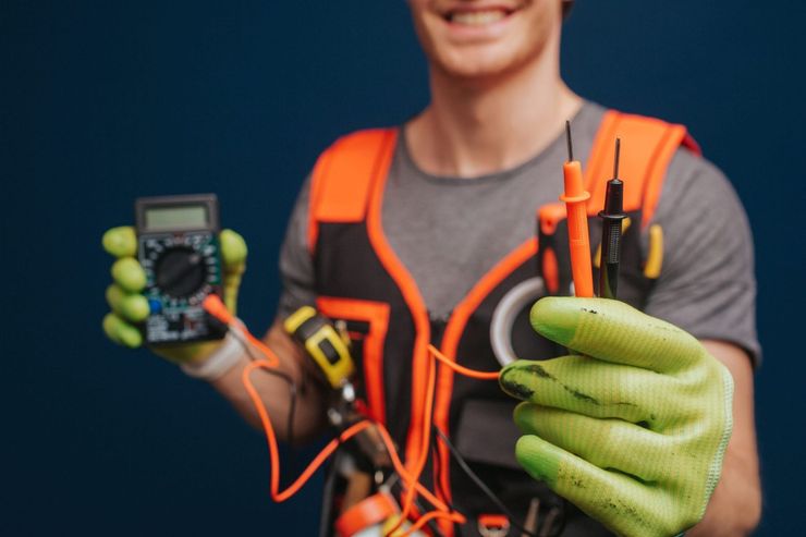 Persona con chaleco de seguridad naranja sosteniendo un comprobador de cables y un manojo de cables sobre un fondo azul.