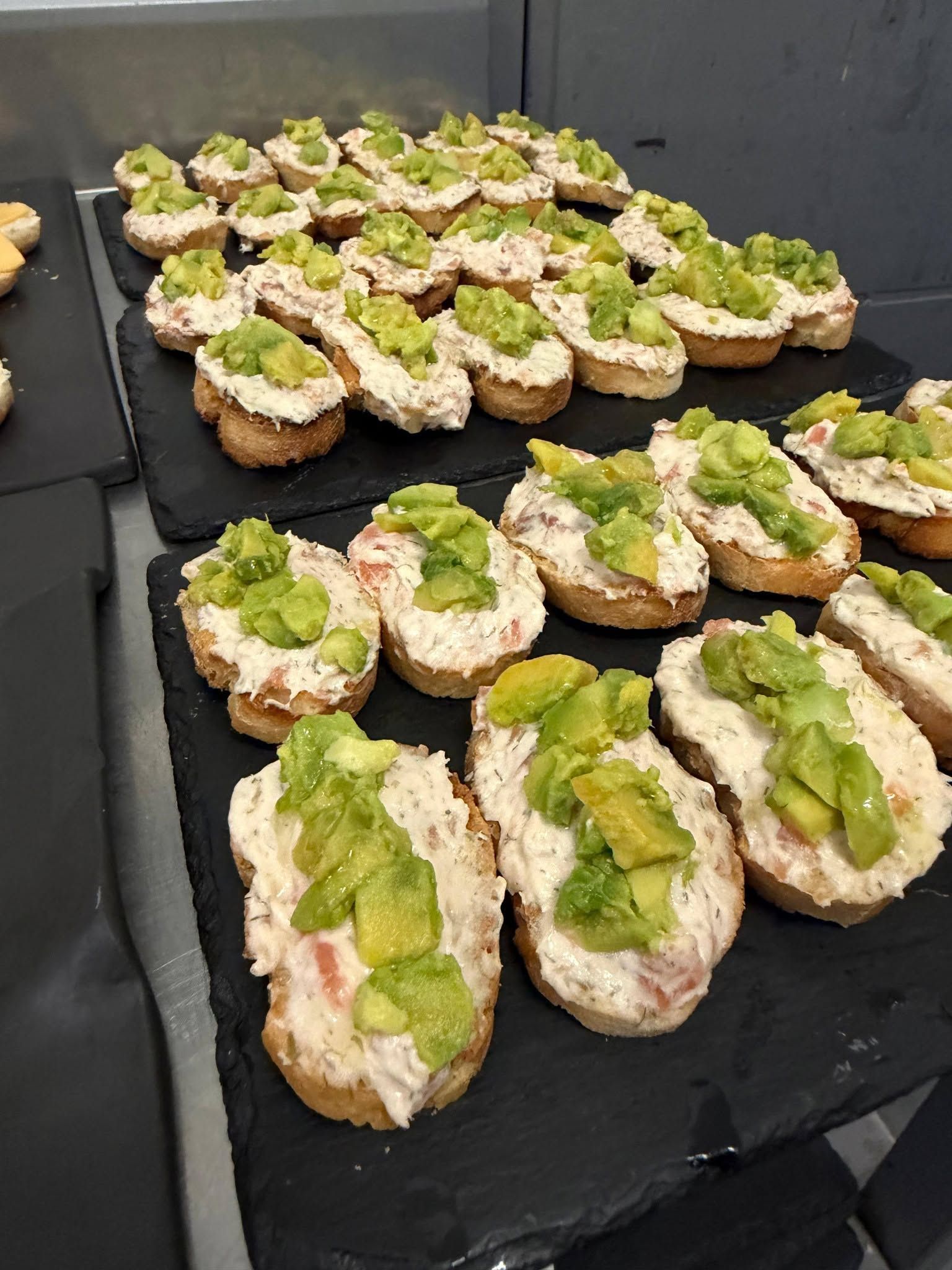 Filas de bocaditos de pan tostado con aguacate en bandejas negras en un evento con servicio de catering