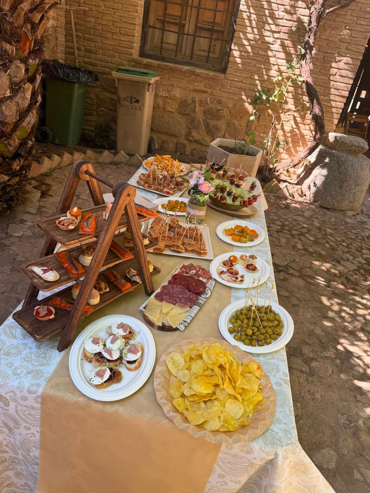Mesa de bufé al aire libre con pasteles, fruta, patatas fritas y salsas en un patio de ladrillo bañado por el sol