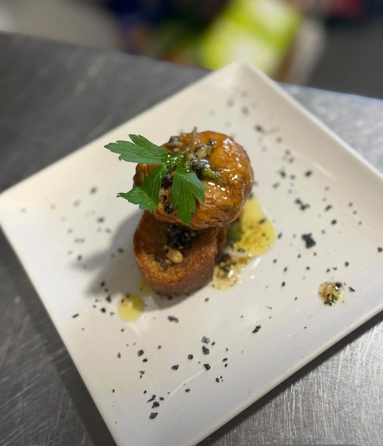 Aperitivo gourmet emplatado, cubierto con perejil y salsa, servido en un plato cuadrado blanco