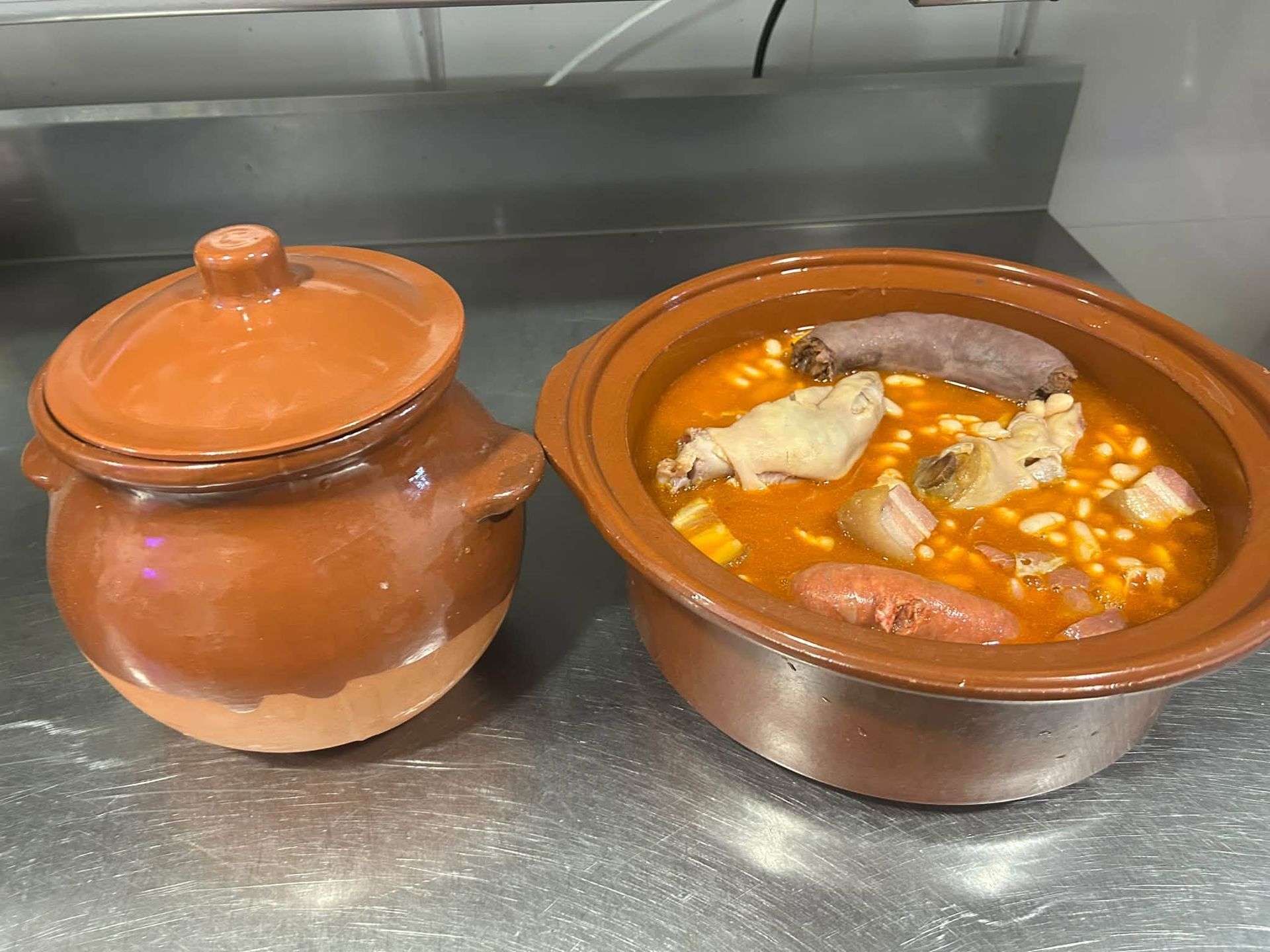 Dos ollas de barro con guiso anaranjado sobre una encimera metálica, una tapada y la otra abierta, con carne y verduras.