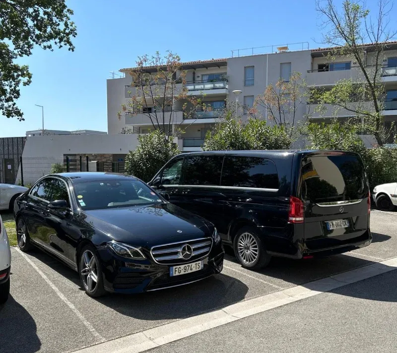 Une berline noire et un monospace noir sont garés sur un parking ensoleillé à côté d'un immeuble d'appartements.