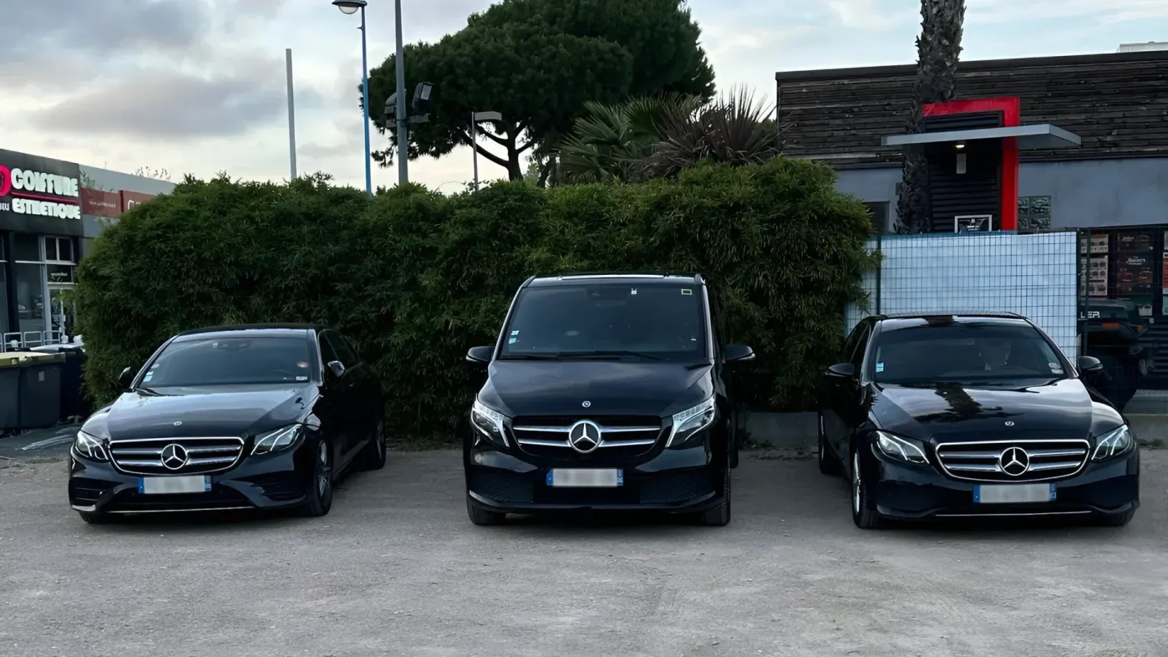 Trois Mercedes-Benz noires garées côte à côte sur un terrain, avec des arbres et des bâtiments en arrière-plan.