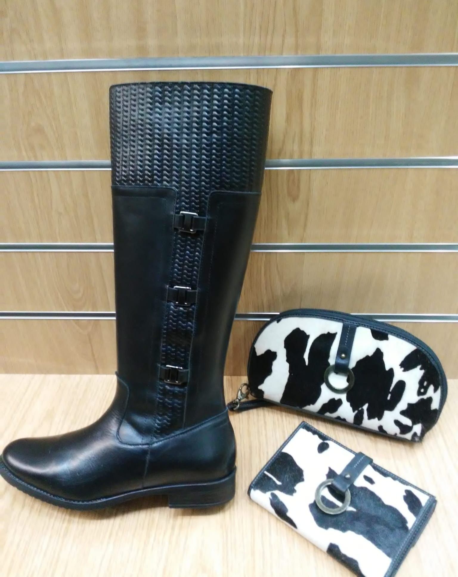 Bota alta de cuero negro hasta la rodilla y dos accesorios con estampado de piel de vaca, expuestos sobre una superficie de madera.
