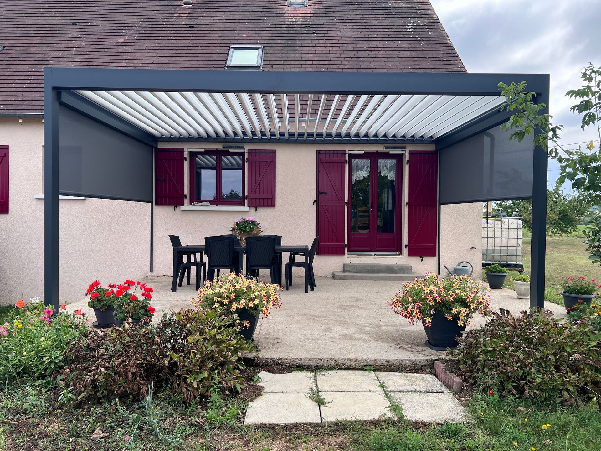 Patio avec une pergola gris foncé, une table et des chaises devant une maison aux volets et à la porte rouges