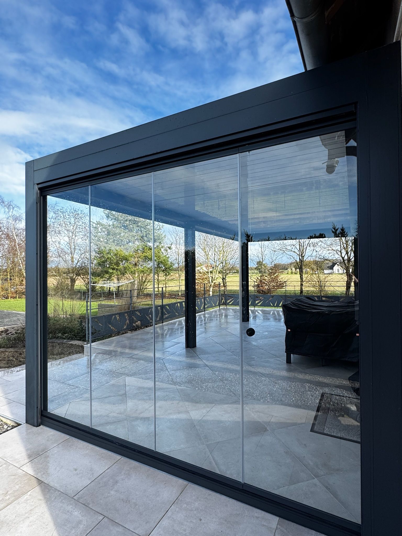 Terrasse vitrée à cadre sombre, donnant sur un paysage sous un ciel bleu