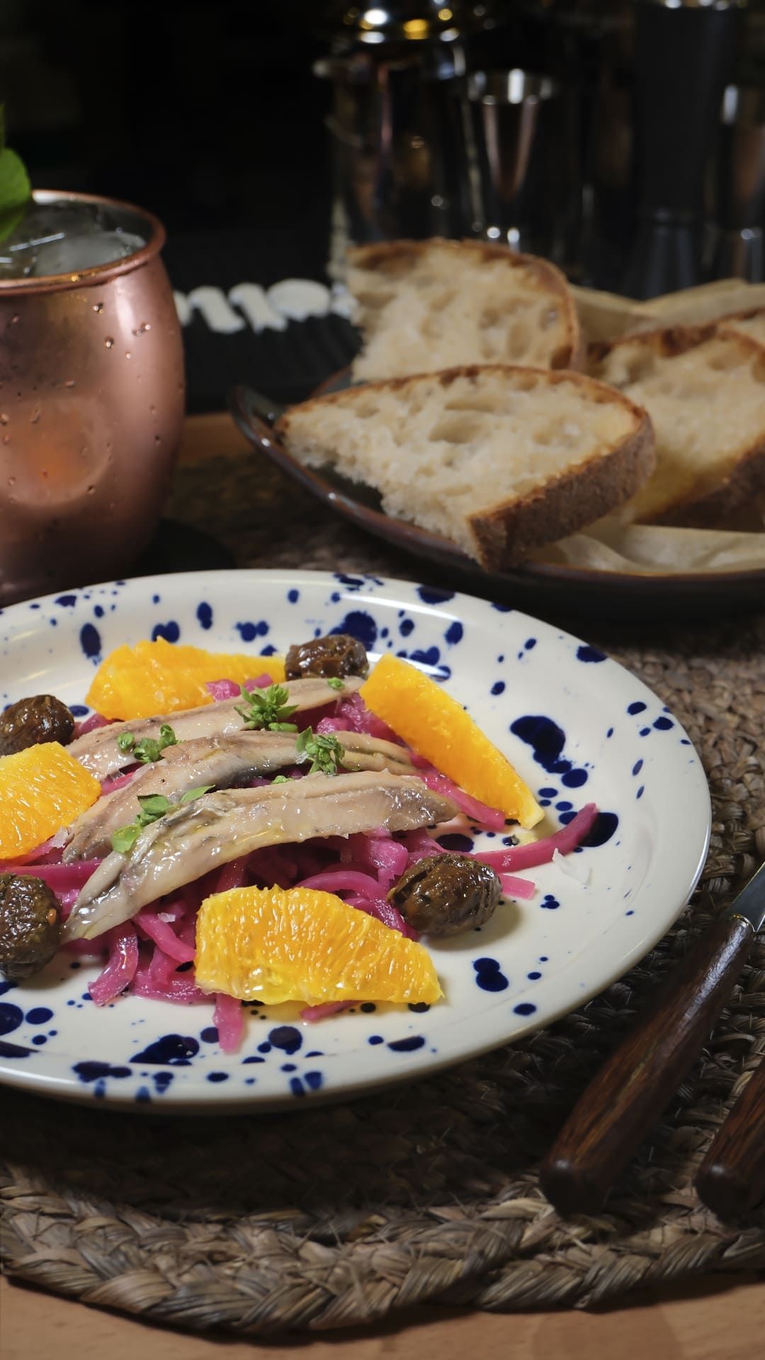 Aperitivo emplatado con rodajas de naranja, verduras y pan sobre una mesa rústica junto a una taza de cobre.