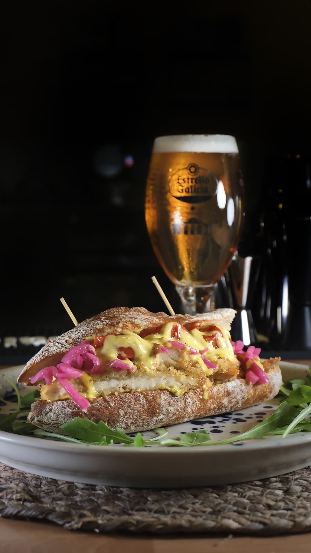 Hamburguesa gourmet con cebollas encurtidas en un plato, servida con una cerveza dorada de fondo.