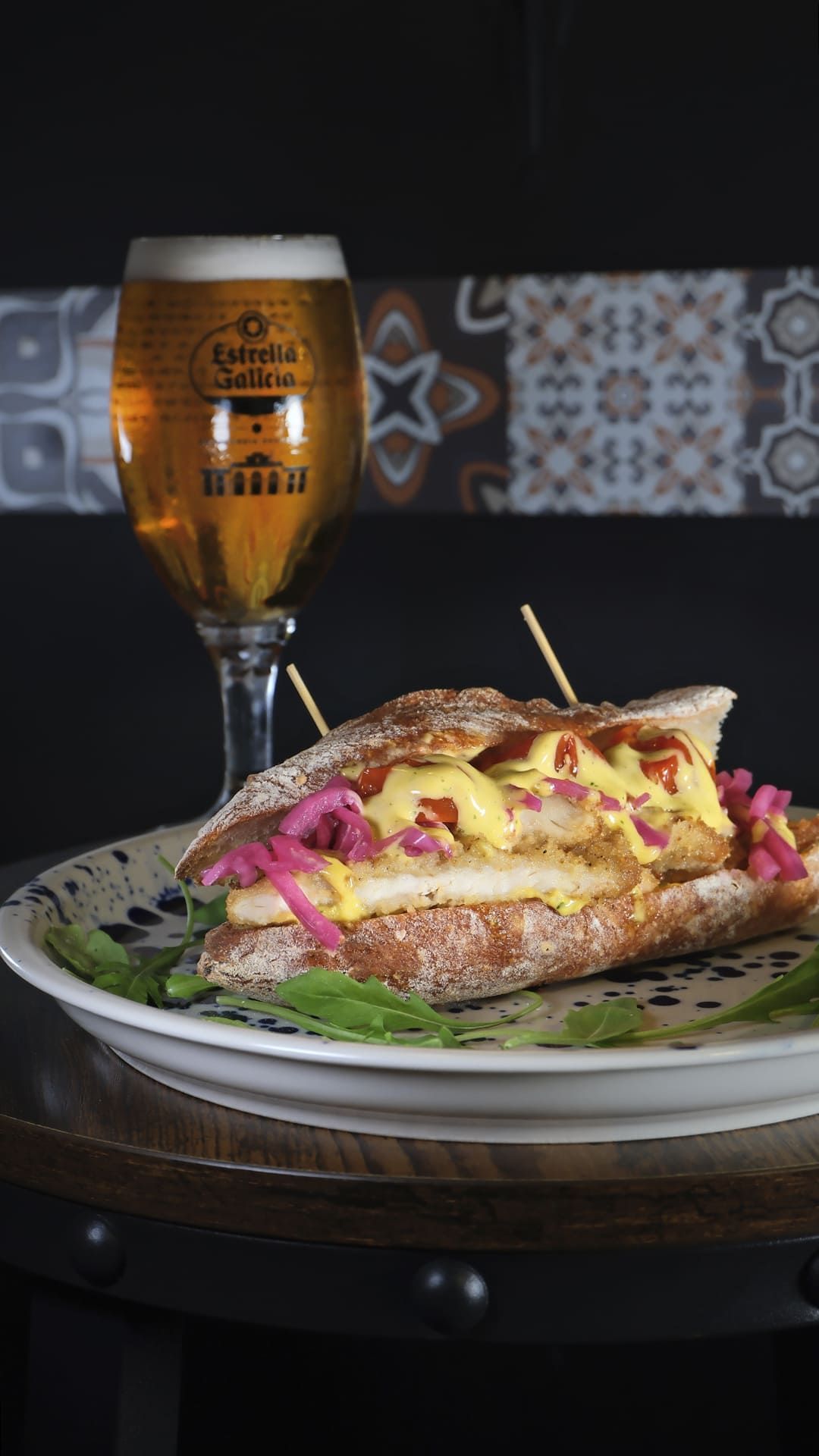 Un vaso de cerveza junto a un sándwich con patatas fritas y salsa sobre una mesa.